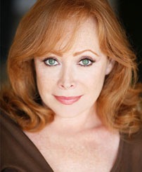A woman with wavy red hair and striking green eyes smiles gently, wearing a brown top. This image highlights her engaging expression and vibrant appearance.