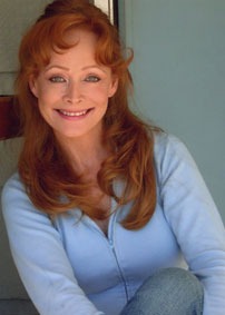 Smiling woman with long, wavy red hair seated casually in a light blue sweater, set against a soft backdrop. Emphasizes warmth and approachability.
