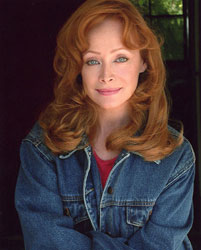 Smiling woman with long, wavy red hair wearing a denim jacket over a red top. The background is softly blurred, creating a warm, inviting atmosphere.
