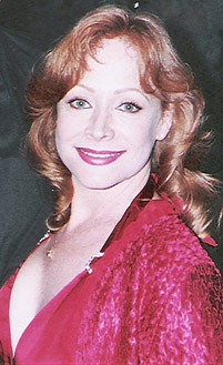 A woman with red curly hair poses in a vibrant red dress, smiling warmly against a dark background, conveying elegance and confidence.